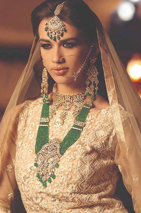 A woman in traditional attire adorned with intricate jewelry, including a green beaded necklace and elegant earrings, with a veil draped over her hair.